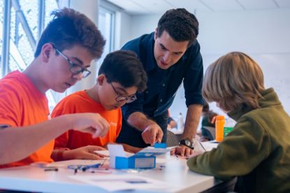 An adult gestures towards the materials on a table as he explains something to three youths working on a hands-on project.