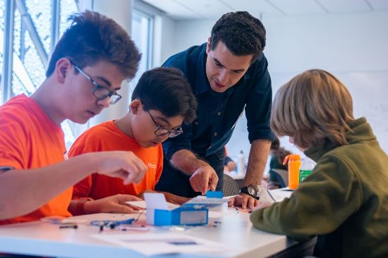 An adult gestures towards the materials on a table as he explains something to three youths working on a hands-on project.