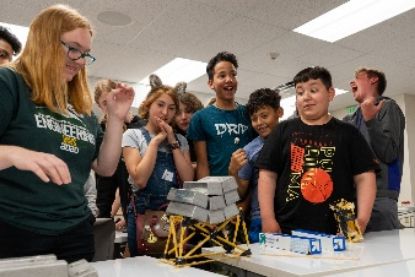 Students surround a table and watch excitedly as weight is added to a model bridge structure.