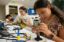 A teen at a lab table scrutinizes a printed circuit board.  Soldering equipment sits on the table before her.  In the background, other teens work on a similar project.