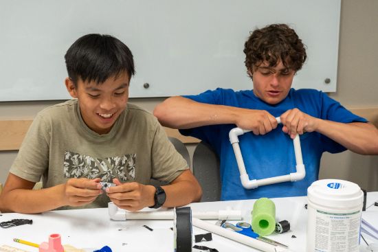 Two students smile as they build a structure with PVC pipe and pool noodle.